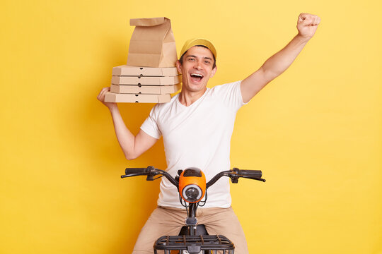 Image of excited delivery man riding bike and holding paper packages with takeaway fastfood, clenched fist, delivering orders fast as super hero, isolated over yellow background.