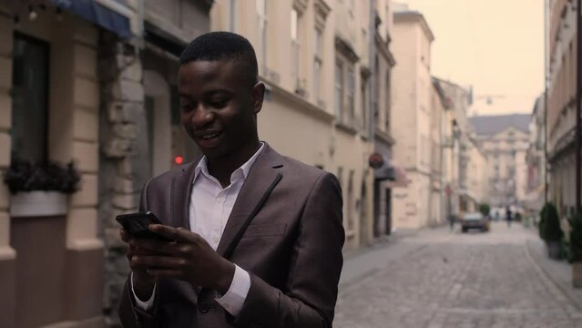 Happy afro american businessman using modern smartphone for working conversation outdoors. Smiling man in suit walking on street and talking on mobile.