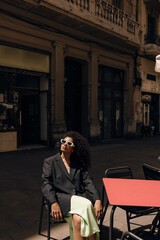 Pretty young african woman sitting in cafe on terrace relaing and enjoying sunny weather. Curly brunette wears dress, black jacket and sunglasses. Concept lifestyle.