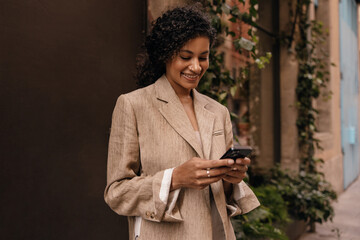 Pretty young african woman texting massages on her smartphone spends time outdoor. Brunette wears light brown jacket. Concept gadgets.