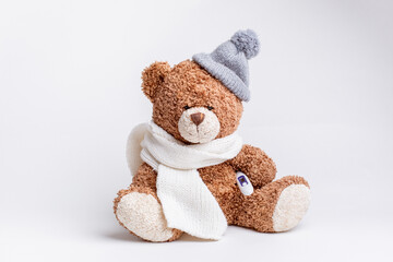 a teddy bear with a thermometer and a knitted scarf and hat is sick on a white background, isolated