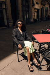 Fototapeta na wymiar Calm young african woman sittin on chair in cafe spends free time outdoor in sunny spring day. Brunette wears dress, jacket and sunglasses. Concept relaxing time.