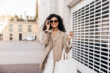 Stylish young african woman in sunglasses stands near backyard city mall looks at camera. Brunette wears classic casual clothes with bag. Concept shopping, weekend.
