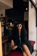 Calm young african woman with curly hair looking at camera, sitting on bench in cafe. Brunette wears green dress and black jacket. Concept fashion, female beauty.