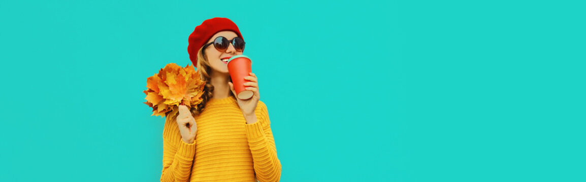 Autumn portrait of happy smiling young woman with yellow maple leaves and cup of coffee wearing knitted sweater, red beret on blue background