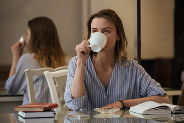 beautiful student girl with long hair drinks coffee in the dining room and reads special educational literature, prepares for classes, exam