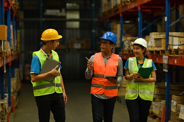 Mature male manager and workers in hardhats and reflective jackets checking quantity of storage product on shelf in a large warehouse