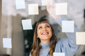 Happy young businesswoman sticking adhesive notes to a glass wall