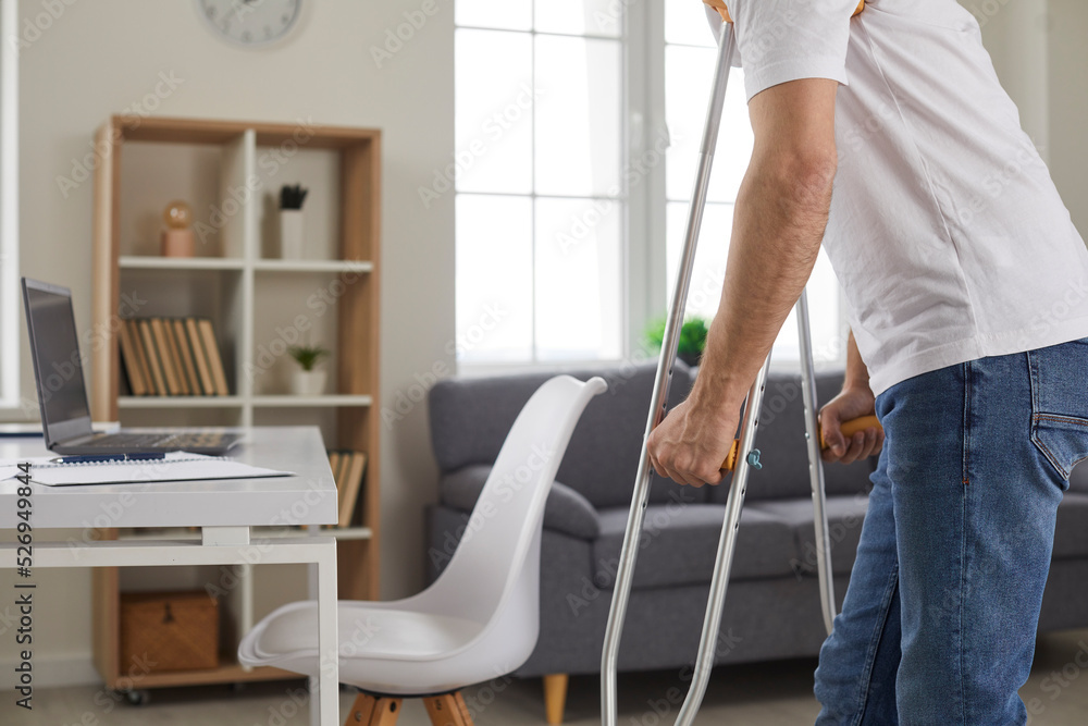 Wall mural Cropped indoor shot of weak young man with disability or plaster cast bandage on broken injured leg using axillary underarm crutches and walking to working desk with laptop computer in home workplace