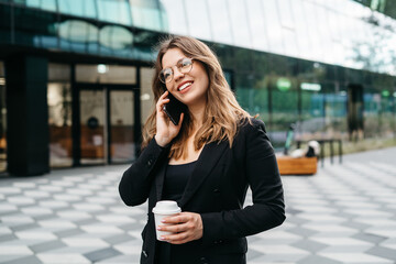 Smiling happy female talking on the smartphone in city downtown, positive woman office employee in smart casual wear chatting on phone on the city street