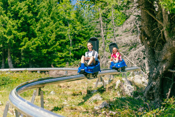 Ski resort mountain coaster. Two girls, one behind the other, are going down the ski slope. Forest and trees in the background - obrazy, fototapety, plakaty