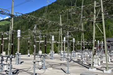 Electric poles with overhead cables, expulsion fuses, surge arrester and other components as a part of hydroelectric power plant on reservoir Mapraggsee. In background is green hill with trees.