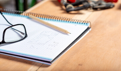 notebook with plan, next to some glasses, on a table, with tools and a pencil.
