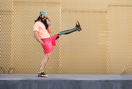 Smiling hipster man with prosthetic leg kicking in front of wall