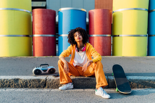 Young woman with skateboard and boom box sitting in front of multi colored pipes