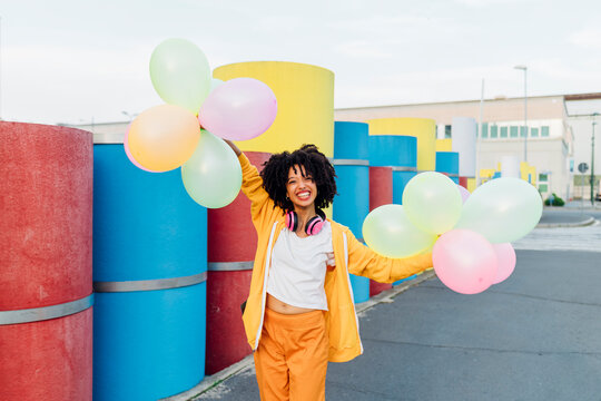 Happy young woman with curly hair holding multi colored balloons on footpath