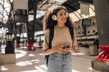 Cheerful young african girl with overhead headphones walks at subway station in sunny weather. Brunette wears t-shirt, jeans skirt and backpack. Concept solo travel, gadgets.