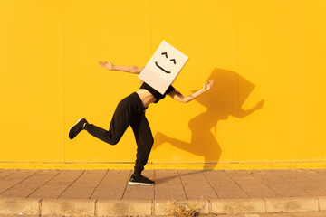 Playful woman wearing box with smiley face doing robot dance in front of yellow wall