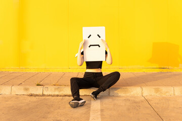Woman wearing box with sad face sitting in front of yellow wall on footpath