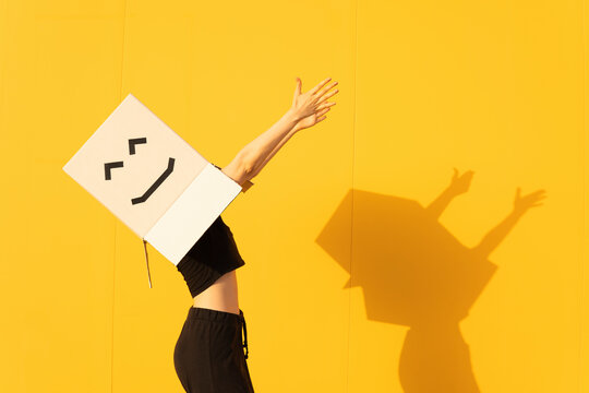 Happy woman wearing box with smiley face dancing by yellow wall