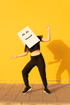 Happy woman wearing box with smiley face doing robot dance in front of of yellow wall on footpath