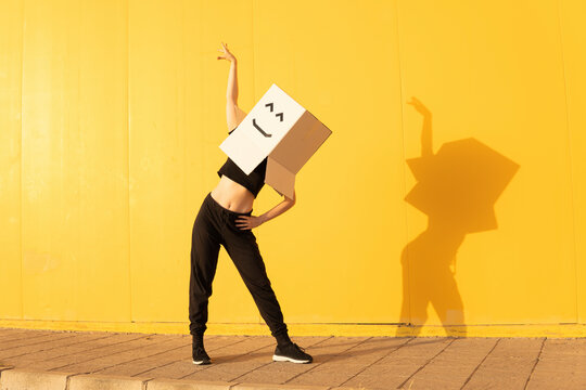 Woman wearing box with smiley face dancing in front of of yellow wall