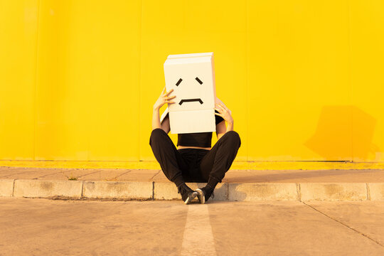 Woman wearing box with sad face sitting in front of yellow wall