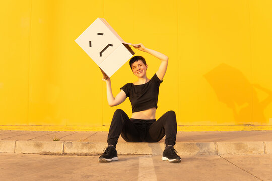 Happy Woman Holding Box With Sad Face Sitting In Front Of Yellow Wall On Footpath