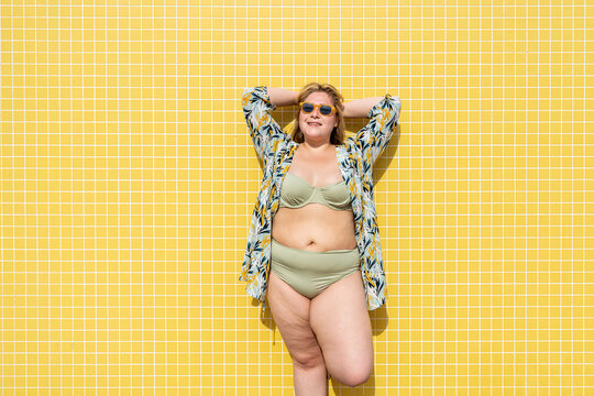 Smiling plus size woman with hands behind head in front of yellow wall