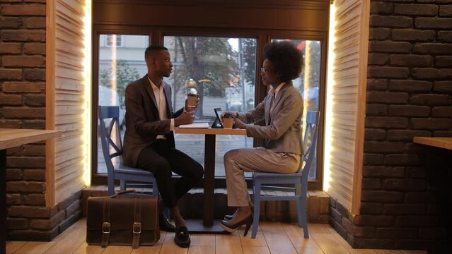 Two young african people in formal clothes sitting at cafe table with digital tablet and documents. Successful businessman and businesswoman cooperating for common project.