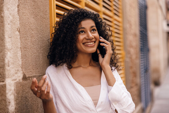 Hopeful Young African Girl With Curly Hair Talking With Family On Phone Outdoor. Model Brunette Hair Wears Singlet And White Shirt. Concept Of Use, Gadgets.
