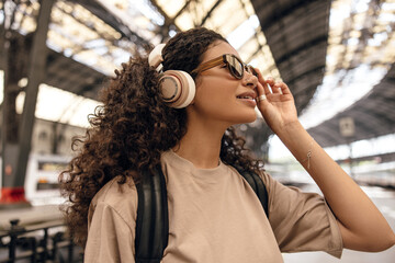 Stylish young african woman with wireless overhead headphones looking away stands on platform alone. Brunette wears light brown t-shirt, sunglasses and backpack. Concept music, travel.