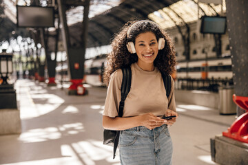Smiling young african woman looking at camera with good mood uses her smartphone on railway station. Brunette curly hair wears casual clothes and backpack. Concept journey.