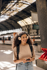 Positive young african woman looking into distance listening music while goes to subway station. Brunette with curly hair wears t-shirt, jeans skirt and backpack. Concept technologies, vacation.