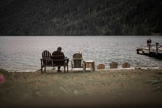 Man sitting on a wooden chair at the beach with a view of a lake and the mountains.