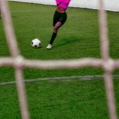 Front view of a unrecognizable dark skinned colored woman in a wrapped up pink purple soccer sports uniform kicking a soccer ball at goal on the football field. Behind the goal view shot.