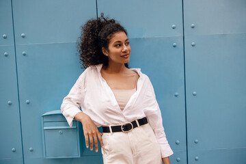Pretty young african woman looking into distance leaned on mailbox gate leading to courtyard. Model...