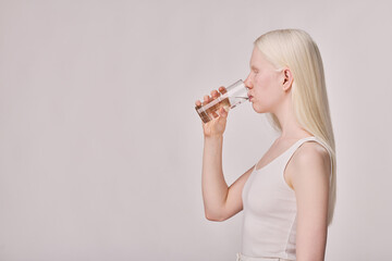 Side view of albino girl with white ong hair drinking water from glass standing on white background - obrazy, fototapety, plakaty