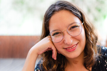 portrait of a young woman with glasses looking at the camera, she is smiling in a blue dress. she is a strong, self-confident and carefree entrepreneur. her hand is touching her face.