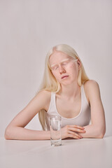 Young albino girl with white long hair sitting at table with glass of water on white background - obrazy, fototapety, plakaty