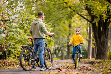 Naklejka premium Happy father and daughter ride bicycles in autumn park on sunny day.