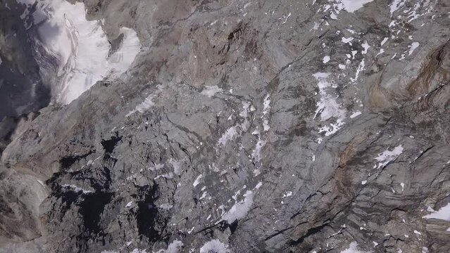 Look down view from drone, pan right, at rugged Alps terrain. Grey rocky landscape with ice and snow pockets.