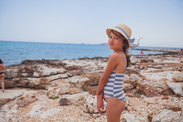 Summer smiling portrait of little girl with straw hat at rocks shore. Adorable child on vacation. Seascape in background. 
