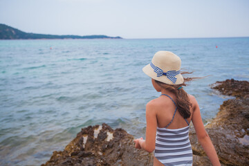 Summer smiling portrait of little girl with straw hat at rocks shore. Adorable child on vacation. Seascape in background. 