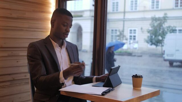 Young african businessman in stylish suit taking notes while looking at tablet screen. Confident man sitting at cafe table and working on distance.