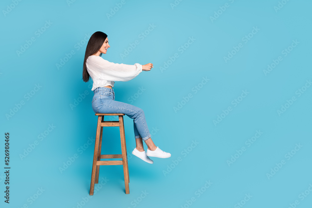 Poster Full size profile photo of positive pretty girl sit stool arms hold imagine wheel look empty space isolated on blue color background