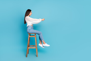 Full size profile photo of positive pretty girl sit stool arms hold imagine wheel look empty space isolated on blue color background
