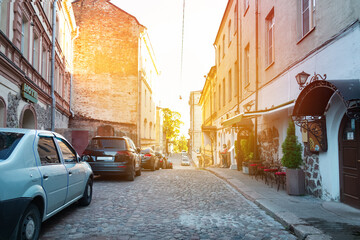 Fototapeta na wymiar Town street with paving stones, houses and parked cars, sunset between buildings