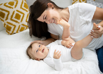 Obraz na płótnie Canvas pretty young and happy mother with dark hair, white t-shirt, blue pants is cuddling in bed with her 7 months old son with blue eyes