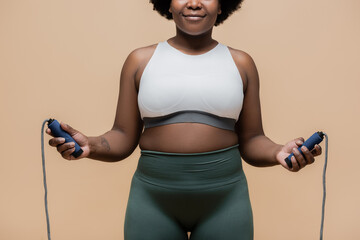 cropped view of cheerful african american plus size woman in sportswear holding skipping rope...